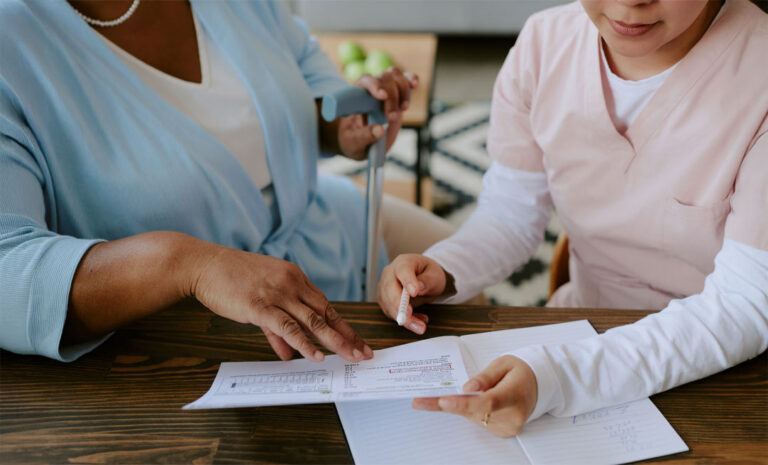Nurse assisting an elderly woman with filling out Medicaid forms at a table, while the woman holds a cane.