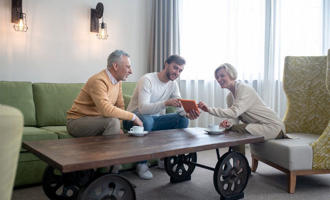 A smiling son visits his parents at their adult day service, sitting together on a couch and looking at photos on a tablet in a bright, comfortable lounge.